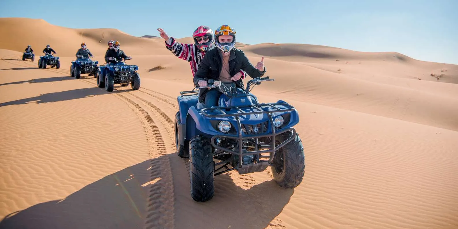 Helmeted students riding a quad bike convoy through Sahara dunes